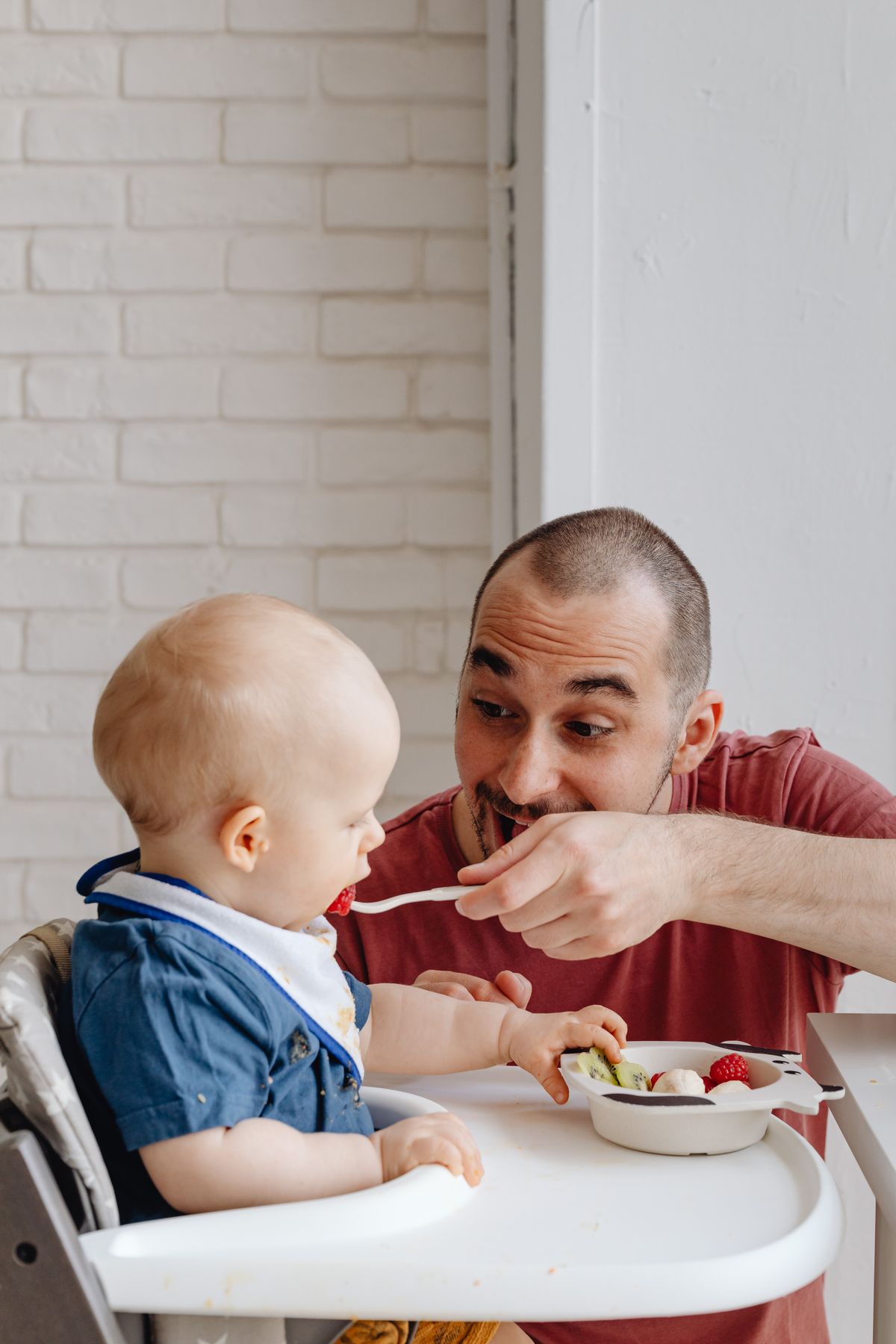 Baby sitting in a high chair eating directly from a bowl on the tray while dad feeds a spoonful