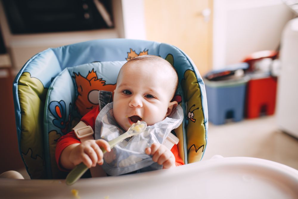 Close-up of a baby eating food with a spoon while sitting in a feeding chair