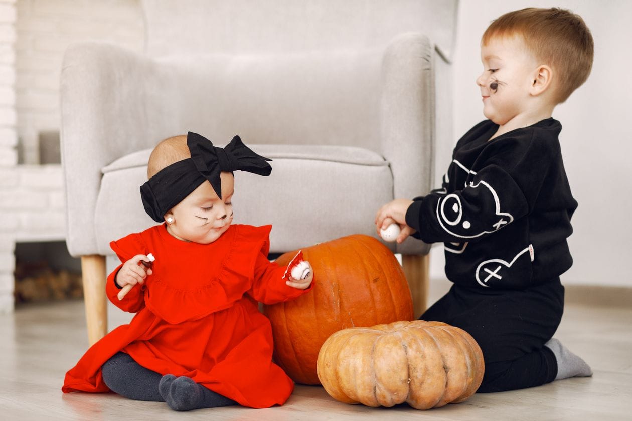 Two young children in halloween costumes playing with pumpkins indoors, an example of hands-on daycare halloween activities for toddlers