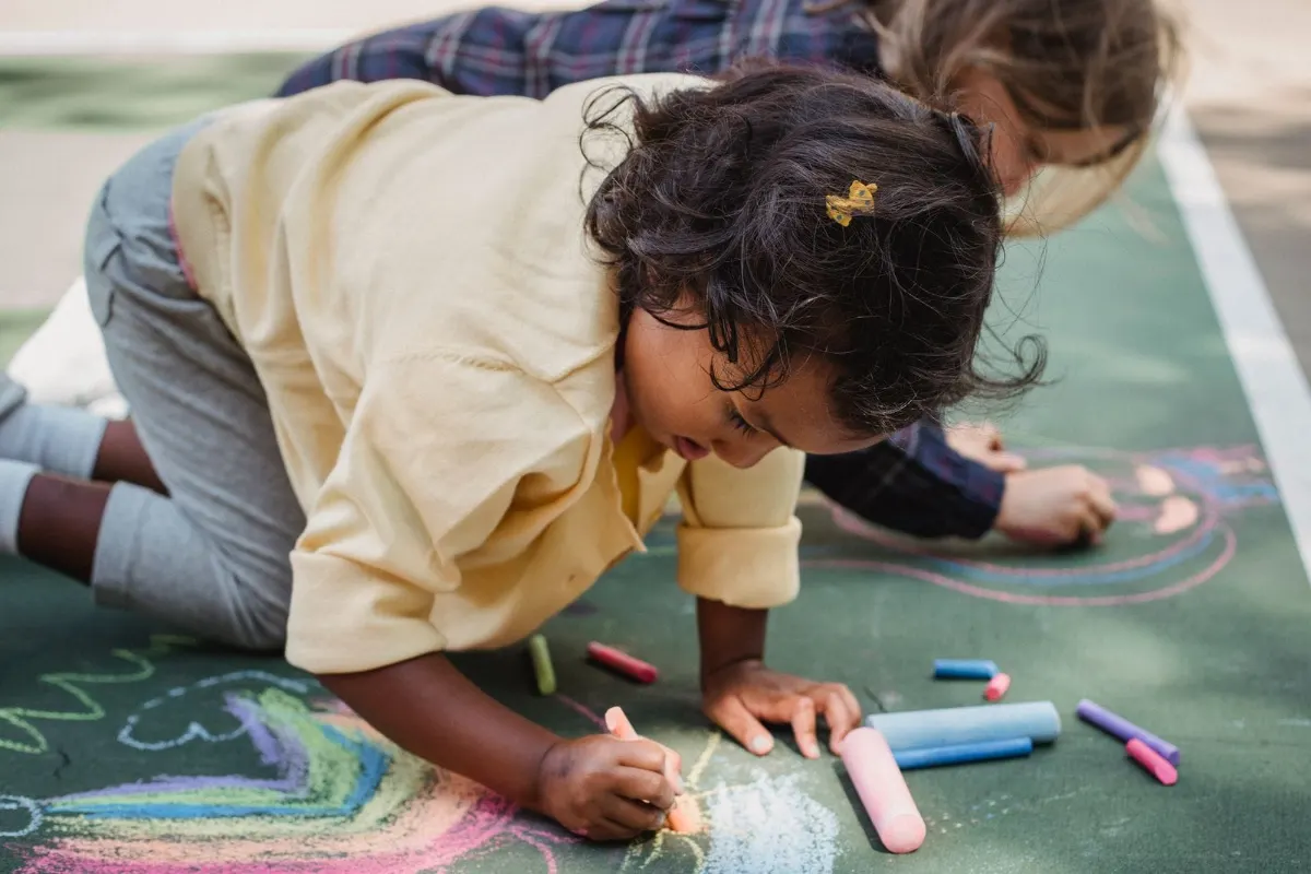 Toddlers drawing with sidewalk chalk on the pavement, part of a chalk hopscotch outdoor activity for toddlers